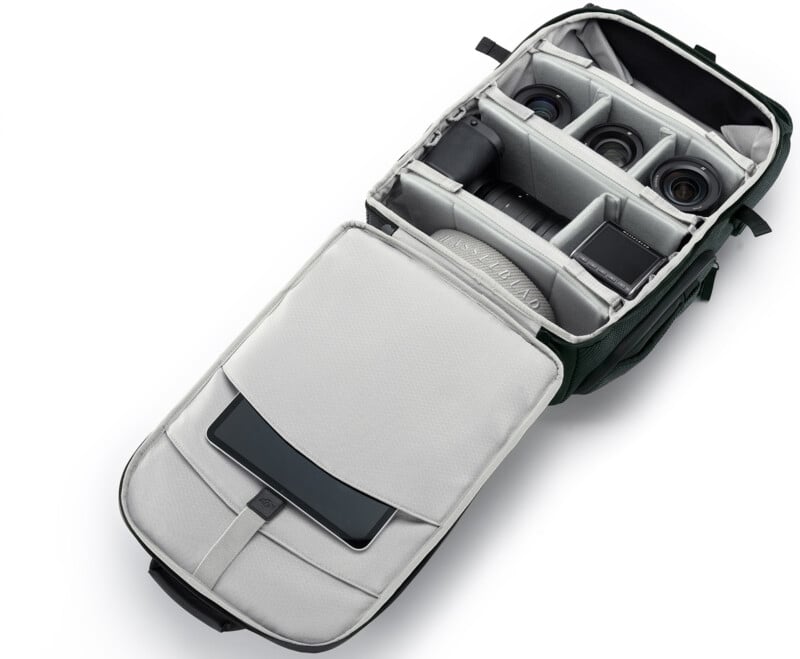 A camera backpack with its main compartment open, showing neatly organized camera gear including a camera body, three lenses, a battery charger, and a tablet stored in an inner pocket.