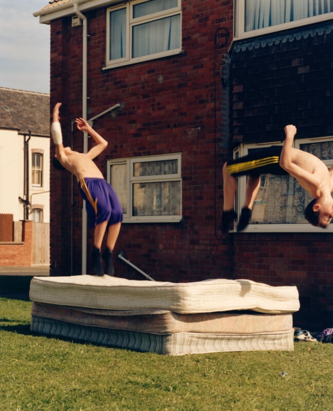 Two shirtless boys wearing athletic shorts perform flips off stacked mattresses on a lawn in front of a brick house with multiple windows, on a sunny day.