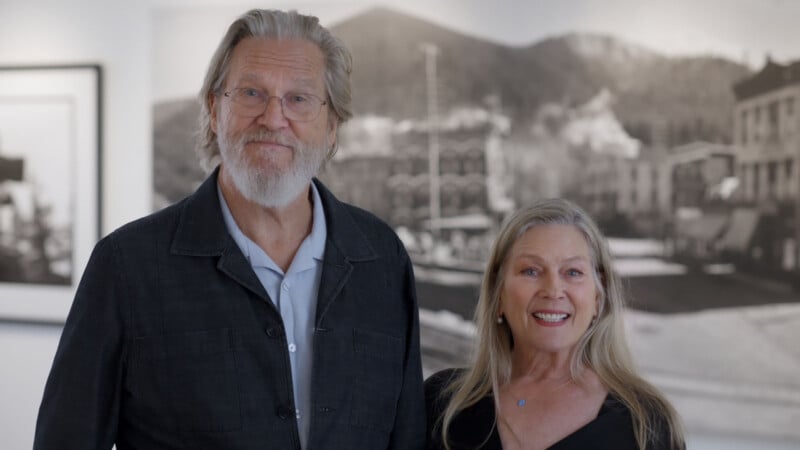 Un hombre mayor con cabello gris y barba se encuentra junto a una mujer sonriente con cabello largo y rubio. Están ubicadas en el interior con un fondo de fotografías borrosas en blanco y negro y vistas a la montaña.
