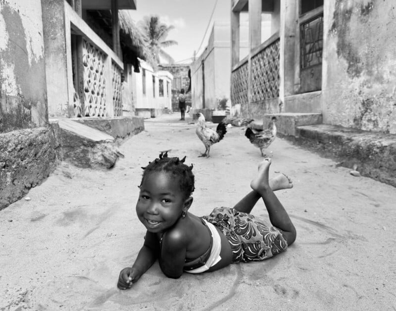 A young girl with braided hair smiles while lying on her stomach in a sandy alley, with chickens walking behind her and worn buildings on both sides. The photo is in black and white.
