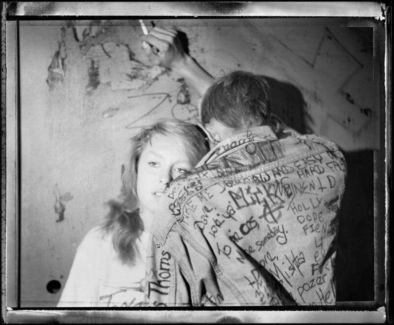 A black-and-white photo of two people standing close together; one faces the camera with a neutral expression, while the other has their back turned, wearing a jacket covered in handwritten graffiti. The wall behind them is marked with scribbles.