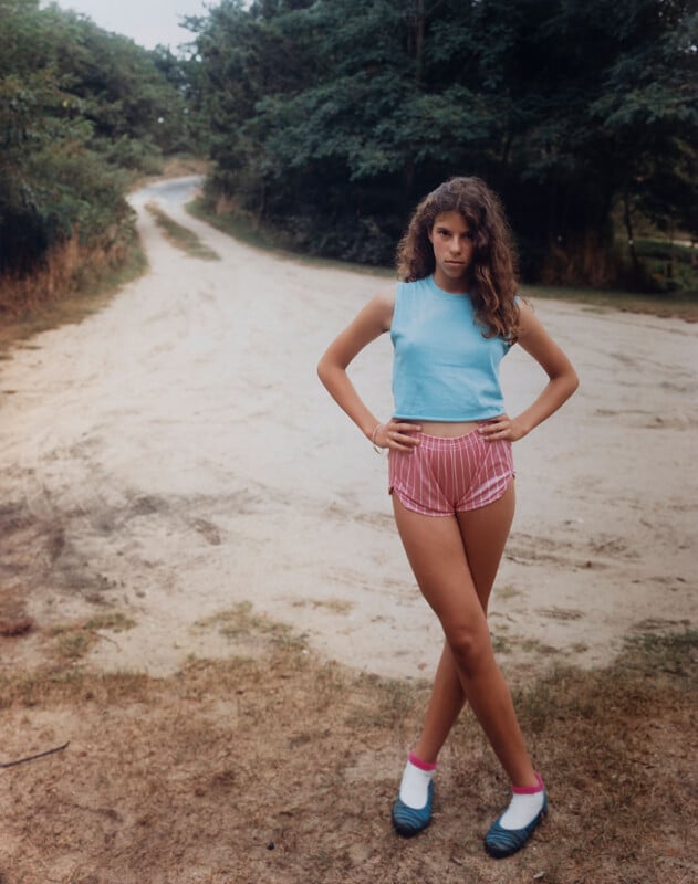 A young woman with curly hair stands on a dirt path in a rural area, wearing a light blue sleeveless top, pink striped shorts, white socks, and blue shoes. Trees and greenery surround the winding path behind her.