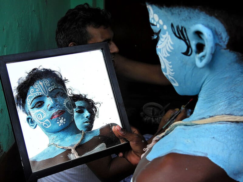 A person with their face and body painted blue and decorated with white designs looks into a mirror, reflecting their painted face while another person is seen in the background.