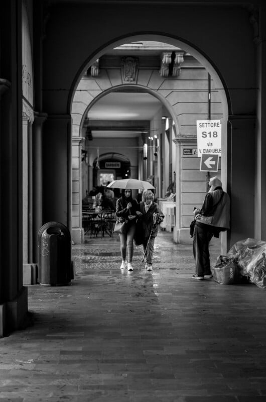 Black and white photo of two people walking under an archway, one holding an umbrella, with a man standing to the right. The street is wet, likely after rain, and there are signs and café tables in the background.