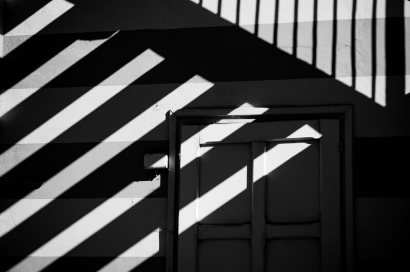 Black and white photo of a closed door on a striped wall, with dramatic diagonal shadows cast by sunlight, creating a pattern of light and dark across the surface.