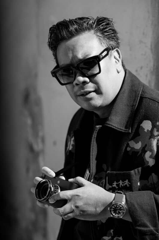 A man with short, styled hair and wearing dark sunglasses looks at the camera while holding a vintage film camera. He is dressed in a patterned jacket and a watch, and the photo is in black and white.