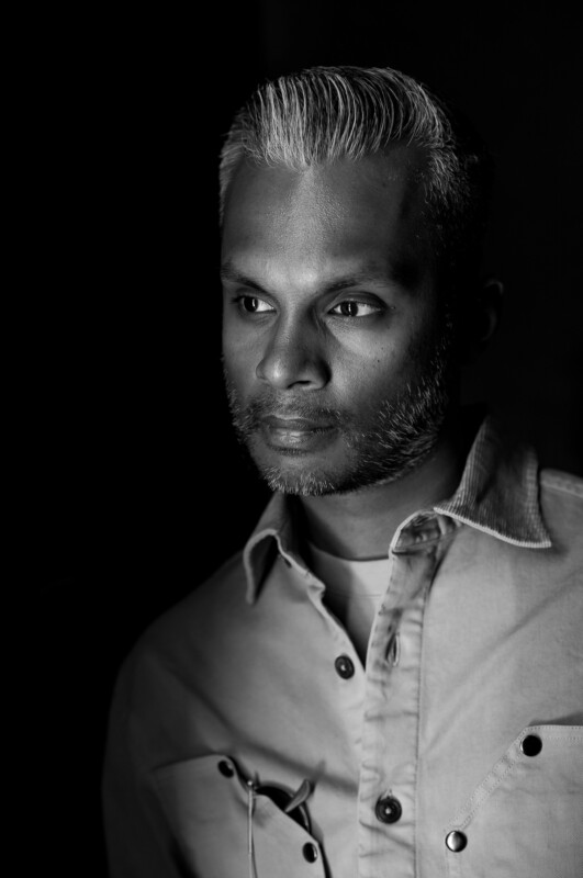 Black and white portrait of a man with short, neatly styled gray hair and a trimmed beard, wearing a button-up shirt and looking thoughtfully to the side. The background is dark, highlighting his face.