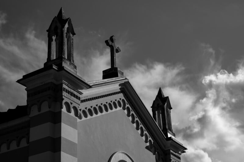 Fotografía en blanco y negro del techo de una iglesia con dos pequeñas agujas y una cruz central, con nubes dispersas en el cielo. El edificio presenta adornos decorativos y detalles arqueados.