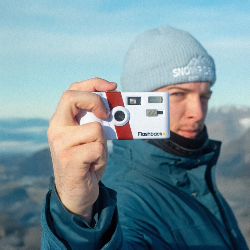 A person in a blue jacket and light blue beanie holds a disposable camera labeled "Flashback" towards the viewer, with mountains and a cloudy sky in the background.