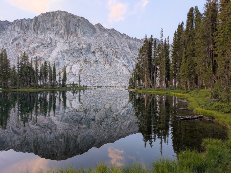 Una montaña con laderas rocosas se refleja en un lago tranquilo al atardecer bajo un cielo parcialmente nublado, rodeado de árboles de hoja perenne y hierba verde.