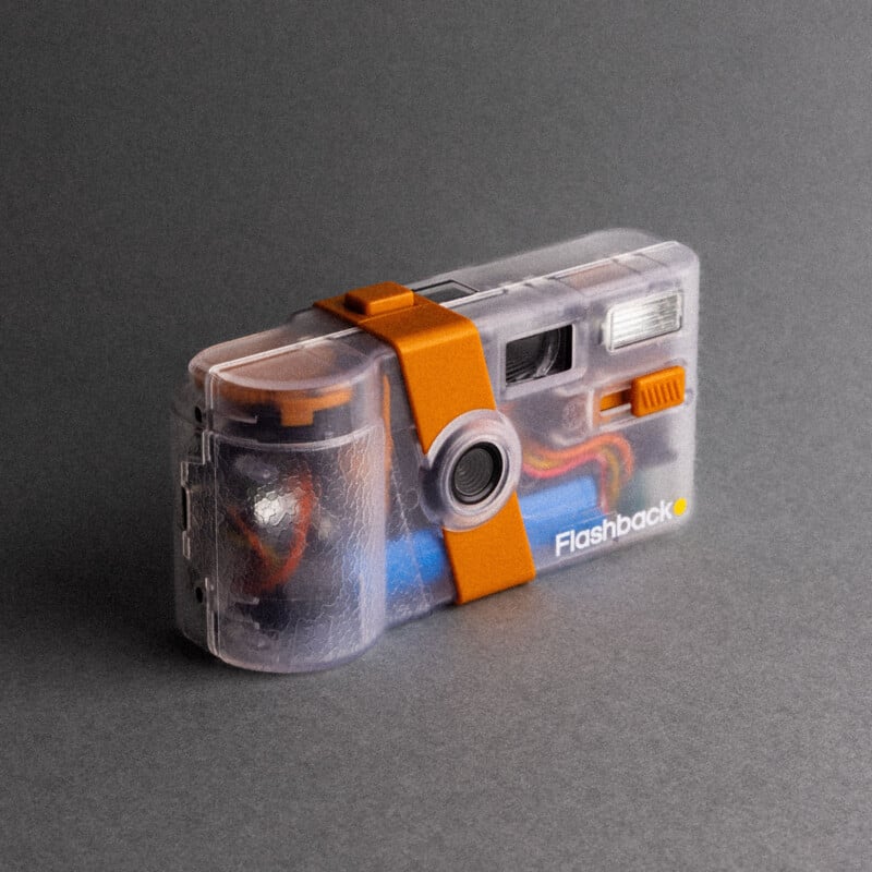 A translucent plastic camera with orange accents and visible internal components, labeled "Flashback," rests on a gray surface against a gray background.