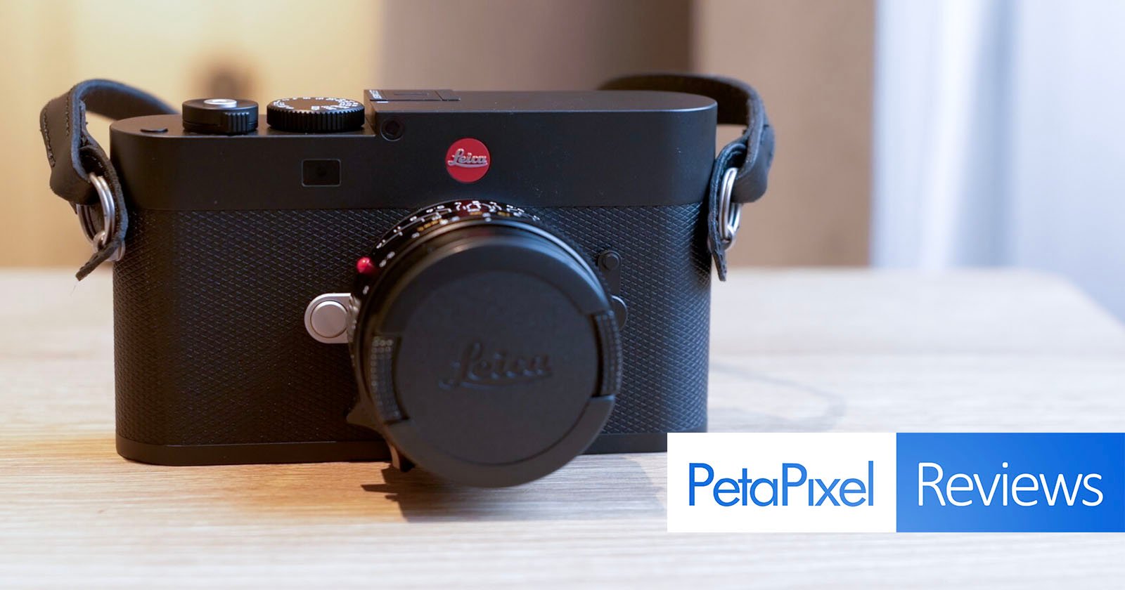 A black Leica camera with a lens cap is placed on a light wooden surface. The image features the "PetaPixel Reviews" logo in the bottom right corner.