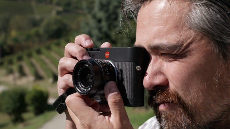 Un hombre barbudo y de pelo gris se encuentra al aire libre, sosteniendo una cámara Leica negra frente a su rostro y mirando a través del visor, con hileras de árboles verdes y viñedos borrosos en el fondo.