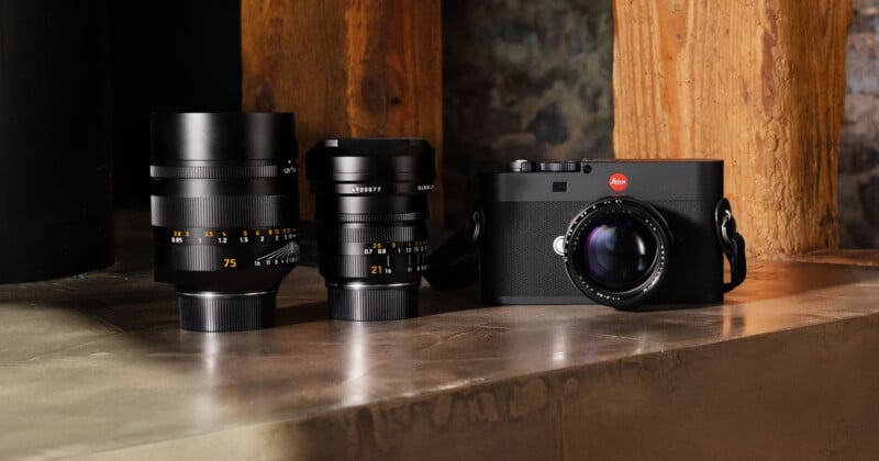 La cámara Leica con lente se muestra sobre una superficie de madera junto a dos lentes de cámara Leica adicionales. El fondo presenta vigas de madera y paredes de piedra.