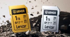 Two Lexar 1TB SD memory cards, one gold and one silver, are standing upright on a rocky surface with debris flying around them. Both cards are labeled "ARMOR" and display speed and capacity details on the front.