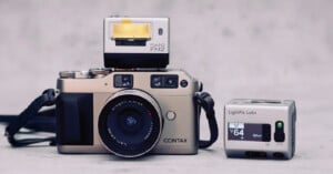 A silver Contax film camera with a lens is placed on a flat surface. A Flash FM2 unit sits on top, and a small LightPix Labs camera accessory is positioned to the right. The background is plain and gray.