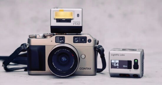 A silver Contax film camera with a lens is placed on a flat surface. A Flash FM2 unit sits on top, and a small LightPix Labs camera accessory is positioned to the right. The background is plain and gray.