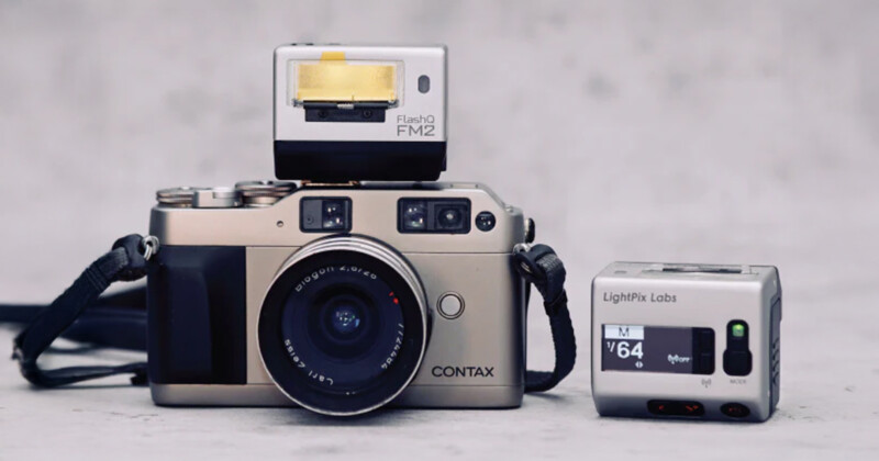 Coloque la cámara de película Contax plateada con la lente sobre una superficie plana. La unidad Flash FM2 está en la parte superior y el pequeño accesorio de cámara LightPix Labs está a la derecha. El fondo es sólido y gris.