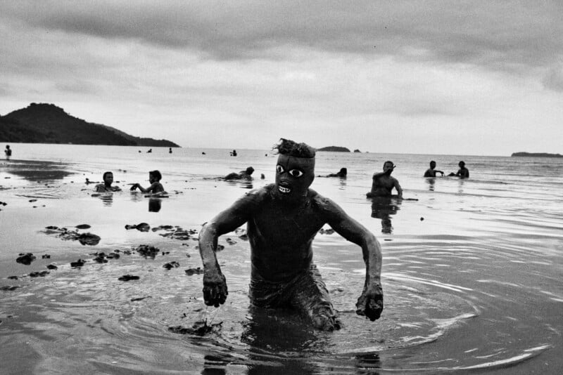 A person covered in mud with a mask and bared teeth emerges from shallow water, with others similarly covered in mud in the background. The scene takes place at a beach under a cloudy sky.