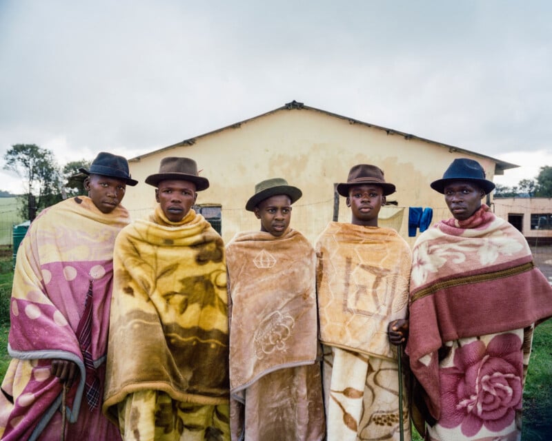 Five young men wearing brimmed hats and patterned blankets stand side by side in front of a beige building, looking toward the camera, with a cloudy sky overhead and grass underfoot.