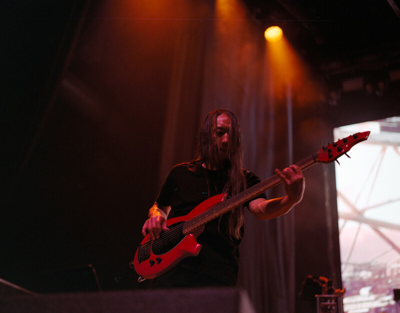 Un músico de pelo largo toca un bajo eléctrico rojo en el escenario bajo espectaculares luces naranjas, con una pantalla al fondo que muestra imágenes abstractas.