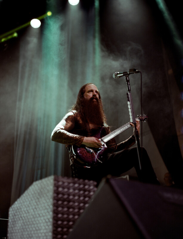 Un hombre barbudo vestido con ropa oscura se sienta en el escenario tocando una guitarra eléctrica bajo espectaculares luces de escenario con humo y un micrófono frente a él.