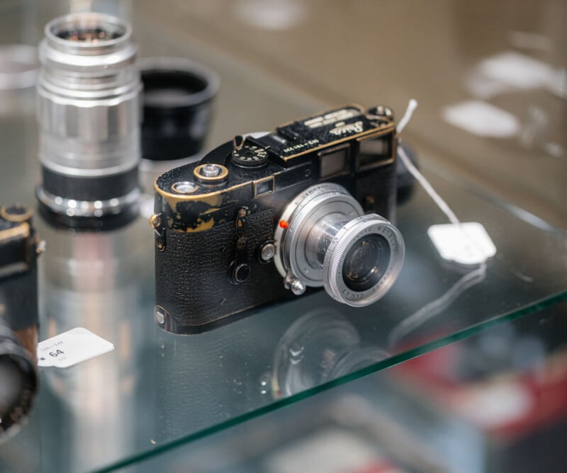 A vintage black Leica camera with a worn finish and silver lens sits on a glass shelf, surrounded by other camera lenses and price tags.