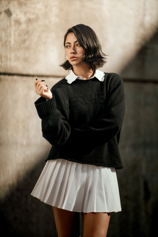 Una mujer con cabello corto y oscuro estaba parada contra una pared texturizada, vestida con un suéter negro, una camisa con cuello blanco y una falda plisada blanca, sosteniendo un bolígrafo o un cigarrillo en la mano, mirando pensativamente hacia un lado.