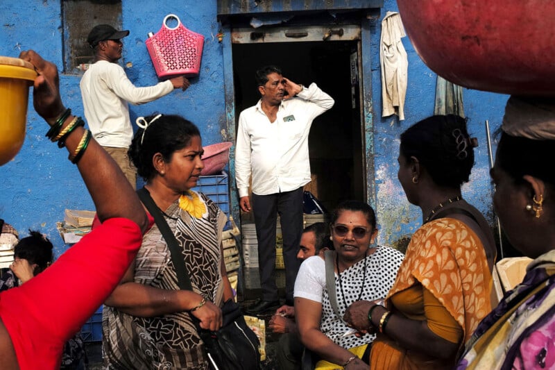 A group of people gathers in front of a blue-painted building. Women in colorful clothing carry items in baskets and tubs. A man in a white shirt stands in the doorway, looking thoughtfully into the distance.