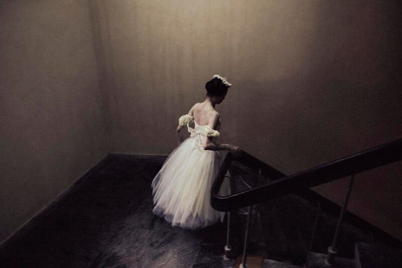 A person in a white, flowing gown and floral headpiece walks up a dimly lit, narrow staircase, casting a soft shadow on the wall in a quiet, atmospheric setting.