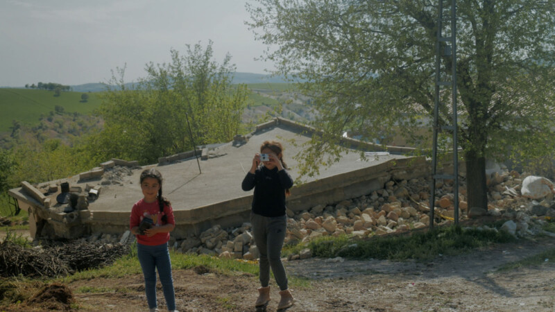 Dos chicas jóvenes se paran en un camino de tierra, una que sostiene una cámara para tomar una foto y la otra sosteniendo un objeto pequeño. Detrás de ellos hay una casa colapsada rodeada de escombros y árboles y colinas verdes en el fondo.