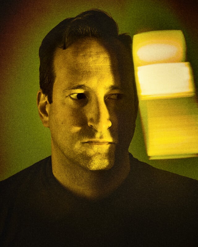 A man with short dark hair looks to the side as a beam of yellow light and shadow falls across his face. The background is green with a blurred, glowing yellow shape, creating a moody, dramatic effect.