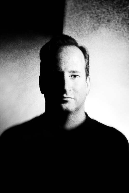 A black-and-white portrait of a man with dramatic lighting that splits his face in half, one side in shadow and the other illuminated, against a textured background.