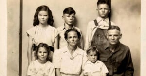 A black-and-white portrait of a family from the early 20th century, showing two adults and four children. The adults are seated in front, with the children standing and sitting around them, all facing the camera with neutral expressions.