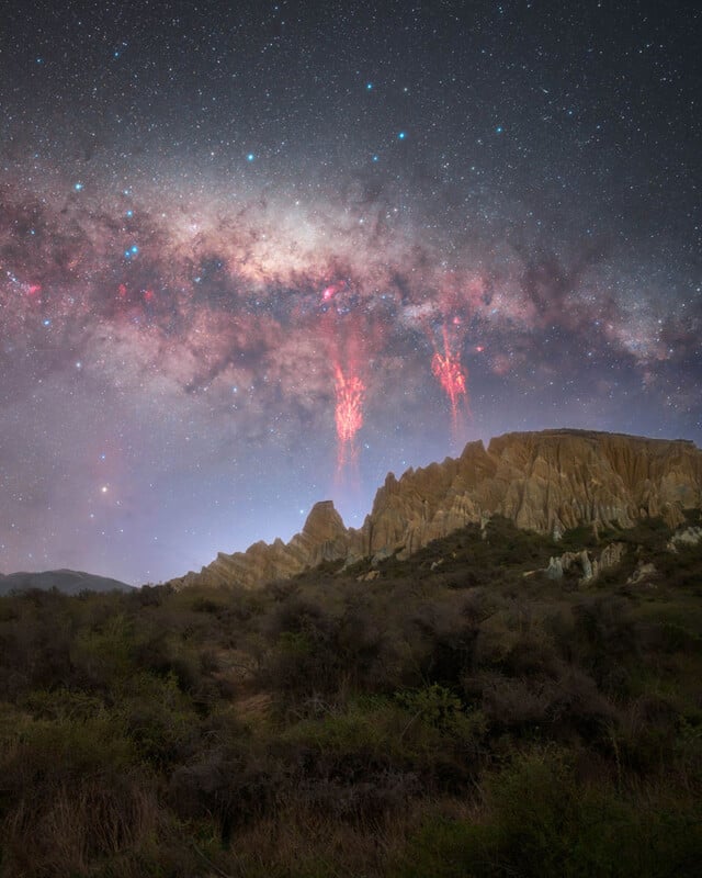 A rugged rocky ridge rises above brushy terrain under a star-filled night sky, with the Milky Way and two bright red lightning-like sprites illuminating the atmosphere above the ridge.