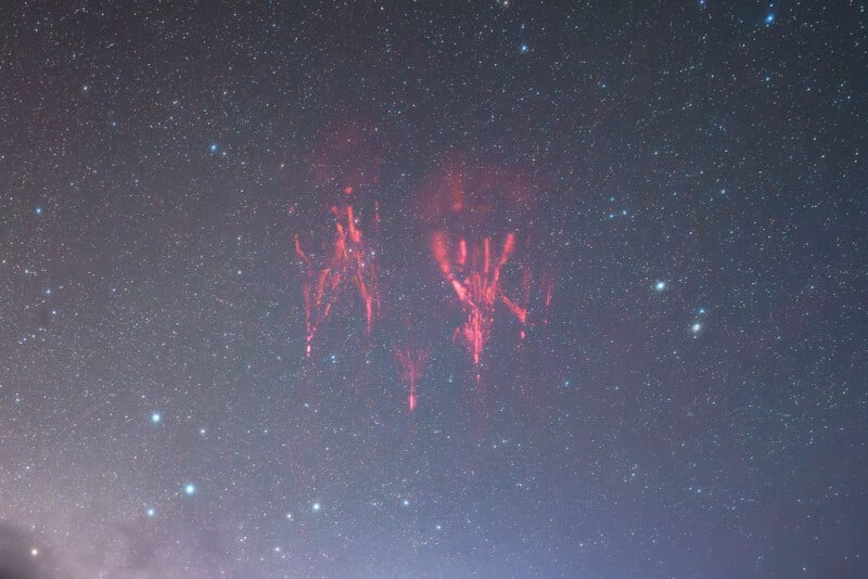 Flashes of red sprites glow faintly in the night sky above a star field, creating trails and tendrils of red light in the upper atmosphere amid a bright, star-filled background.