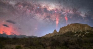 A rocky desert landscape under a starry night sky with the Milky Way visible. Red streaks of light appear in the sky above jagged cliffs, and a haze of orange clouds glows on the horizon. Shrubs cover the foreground.