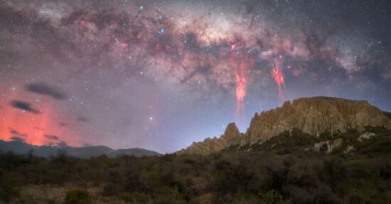 A rocky desert landscape under a starry night sky with the Milky Way visible. Red streaks of light appear in the sky above jagged cliffs, and a haze of orange clouds glows on the horizon. Shrubs cover the foreground.