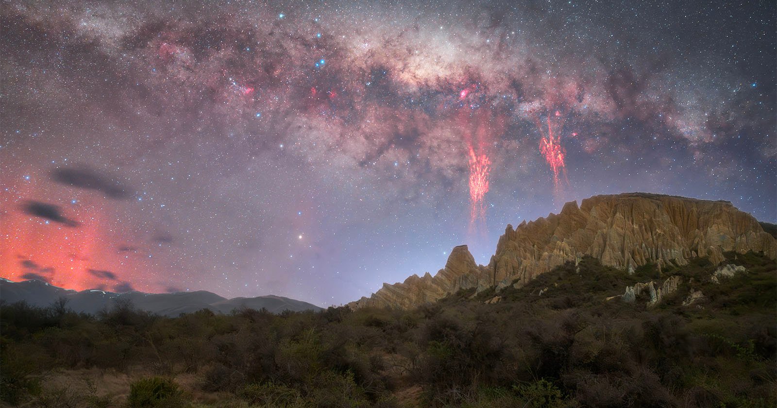 A rocky desert landscape under a starry night sky with the Milky Way visible. Red streaks of light appear in the sky above jagged cliffs, and a haze of orange clouds glows on the horizon. Shrubs cover the foreground.