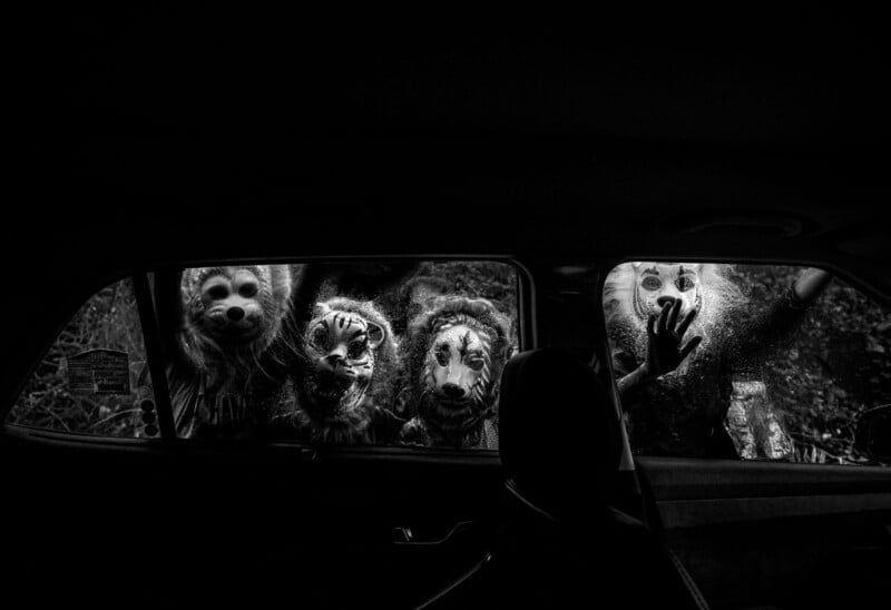 Four people in animal masks peer into a car window from outside, their faces eerily lit. The photo is black and white, with a dark interior and dense foliage visible behind the figures.