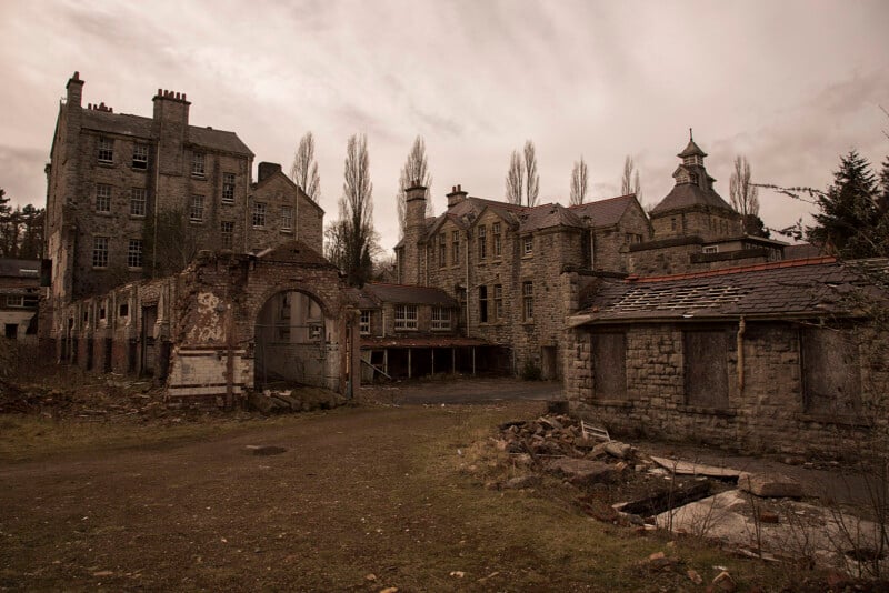 An abandoned stone building complex with crumbling walls, broken windows, and overgrown vegetation under a cloudy, overcast sky, giving a sense of decay and neglect.