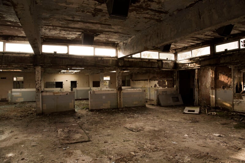 Abandoned, decaying building interior with broken windows, peeling paint, and debris scattered on the floor. Large square columns and tiled partitions divide the space, with natural light streaming through the windows.