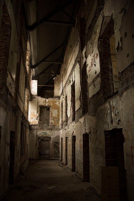 A dark, abandoned building interior with peeling walls, exposed brick, debris on the floor, and missing windows, lit dimly by natural light coming from an opening above.