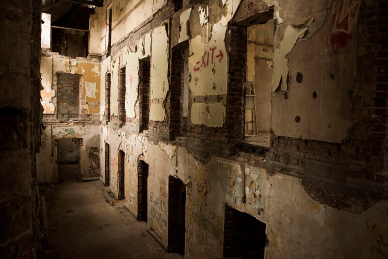 A dimly lit hallway inside an abandoned, decaying building with exposed brick, peeling paint, missing doors and windows, and graffiti on the damaged walls. Light enters through gaps in the ceiling above.