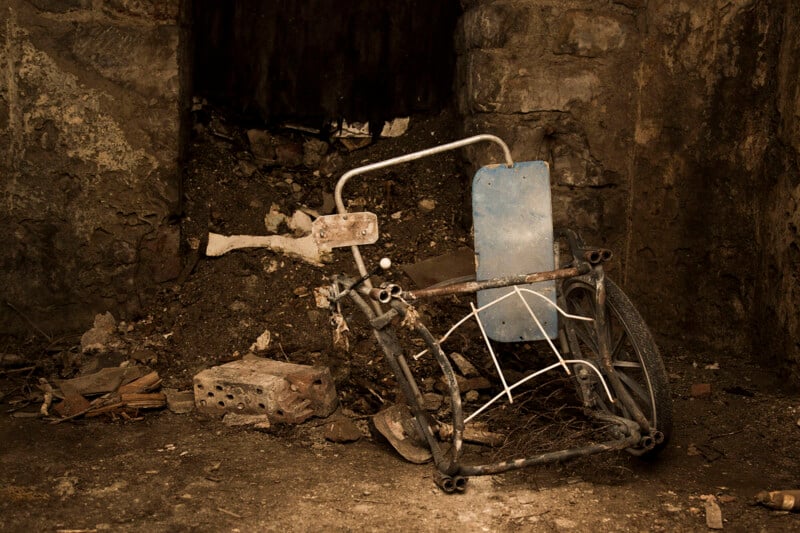 A broken, rusted wheelchair with a blue backrest sits abandoned in a dim, decaying stone room with dirt, debris, and scattered bricks on the floor.