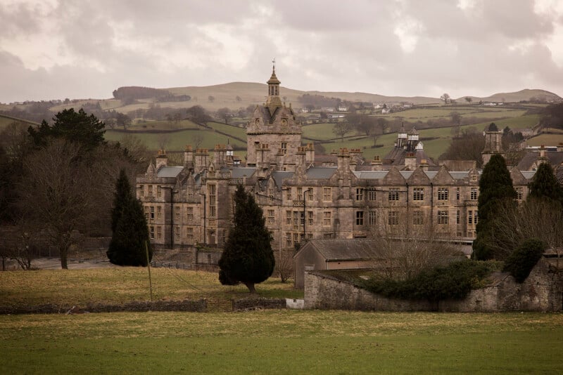 A large, old stone building with a central clock tower stands among trees and grassy fields, with rolling hills and scattered houses in the background under a cloudy sky.
