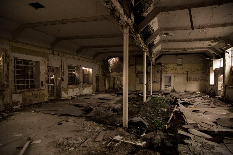 An abandoned, decaying building interior with peeling paint, broken windows, exposed beams, debris, and rubble scattered across the floor. Sunlight filters through damaged sections of the ceiling.
