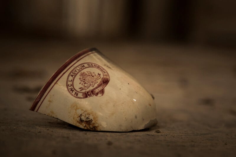 A broken ceramic cup fragment lies on a dusty surface, displaying a maroon logo and the words "NYC Asylum Dendgh" along with a crest-like emblem. The background is blurred and brown-toned.