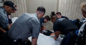 A group of emergency responders surrounds and assists a person lying on a stretcher with a neck brace, preparing to move them in a hallway with beige tiled walls.
