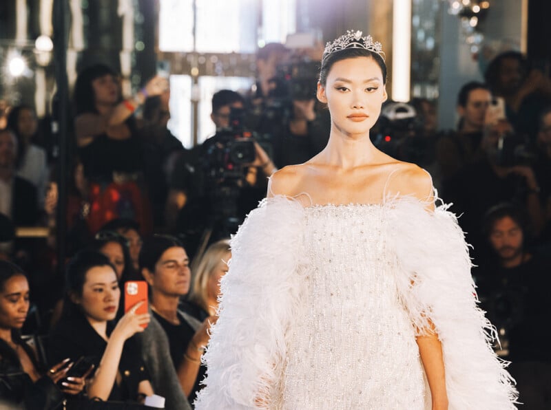 A model wearing a white, feathered gown and a tiara walks the runway at a fashion show, with photographers and audience members taking photos in the background.